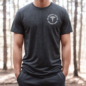 May include: A dark heather grey t-shirt with a white graphic of a medical caduceus symbol and the text "Emergency Nurse" on a circle around the symbol.