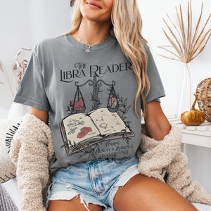 Pu&ograve; includere: Una maglietta grigia con una grafica di un libro e una bilancia, con il testo "The Libra Reader" e "Finding beauty & balance on every page".