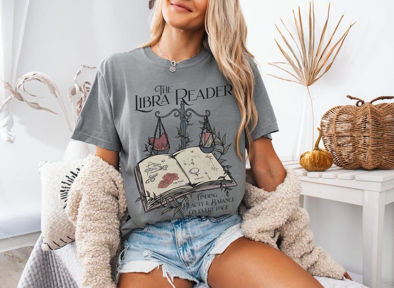 Pu&ograve; includere: Una maglietta grigia con una grafica di un libro e una bilancia, con il testo "The Libra Reader" e "Finding beauty & balance on every page".
