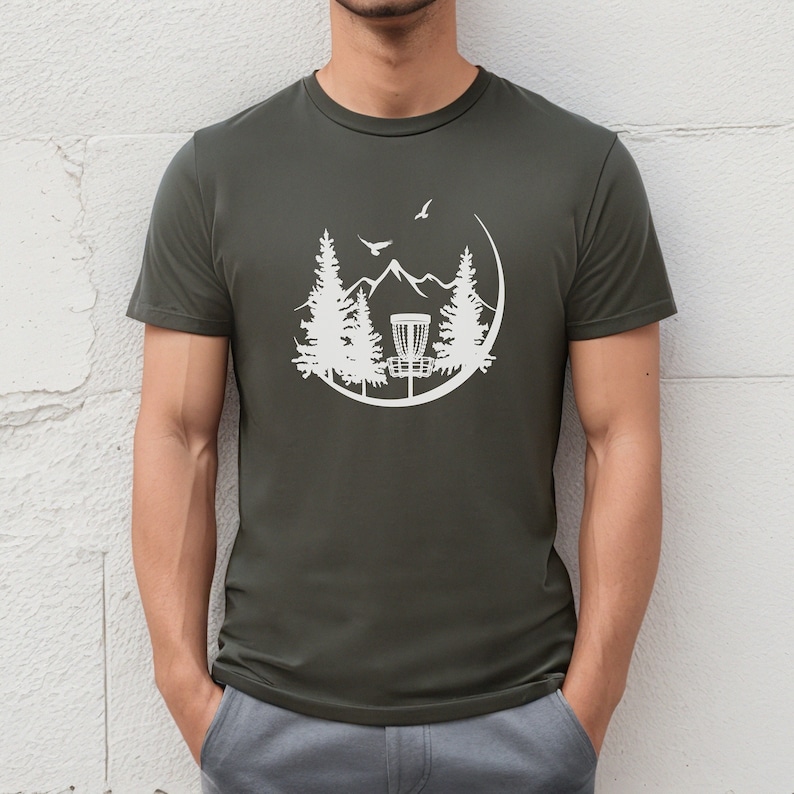 May include: A dark green t-shirt featuring a white graphic of a disc golf basket within a circle, surrounded by trees and mountains. Two birds are depicted flying in the sky.