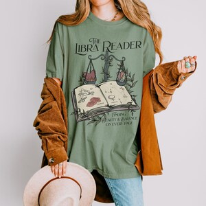 Pu&ograve; includere: Maglietta verde con una grafica di un libro, una bilancia e stelle. Il testo sulla maglietta recita "The Libra Reader" e "Finding Beauty & Balance on Every Page".