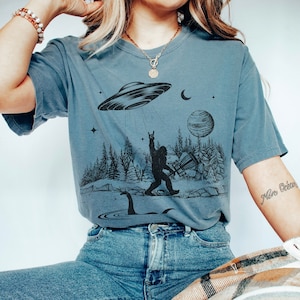 May include: A blue t-shirt featuring a graphic print of Bigfoot playing a guitar, a Loch Ness Monster, a UFO, a crescent moon, a planet, and stars. The shirt has a relaxed fit and short sleeves.