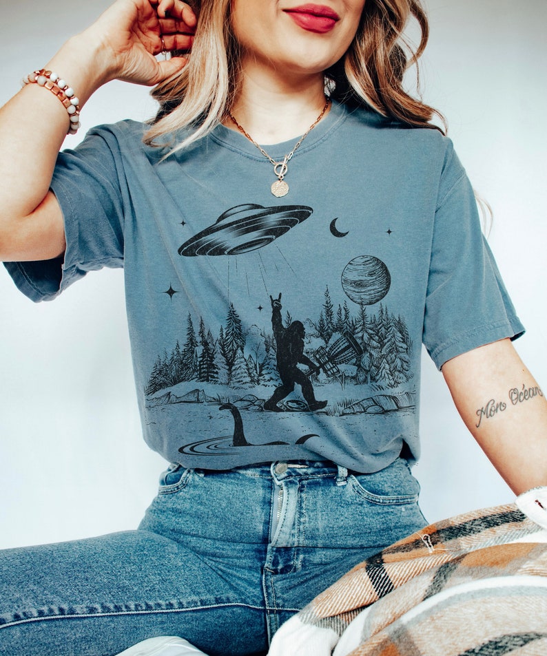 May include: A blue t-shirt featuring a graphic print of Bigfoot playing a guitar, a Loch Ness Monster, a UFO, a crescent moon, a planet, and stars. The shirt has a relaxed fit and short sleeves.