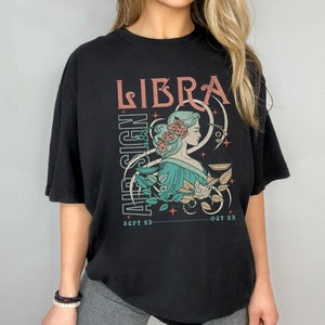 Op de afbeelding: Zwart T-shirt met een grafische print van een vrouw met een bloemenkrans en een weegschaal, met de tekst "LIBRA AIR SIGN SEPT 25 - OCT 23".