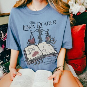 Pu&ograve; includere: Una maglietta blu con un design grafico di un libro, una bilancia e il testo "The Libra Reader: Finding Beauty & Balance on Every Page".