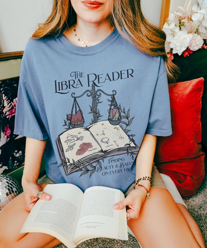Pu&ograve; includere: Una maglietta blu con un design grafico di un libro, una bilancia e il testo "The Libra Reader: Finding Beauty & Balance on Every Page".