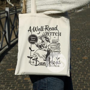 K&ouml;nnte beinhalten: Eine wei&szlig;e Einkaufstasche mit einer Schwarzwei&szlig;illustration einer Hexe, die ein Buch liest. Der Text auf der Tasche lautet "A Well-Read Witch" und "Salem's Finest Witches!" mit einem Stapel B&uuml;cher und dem Text "is a Dangerous Creature".