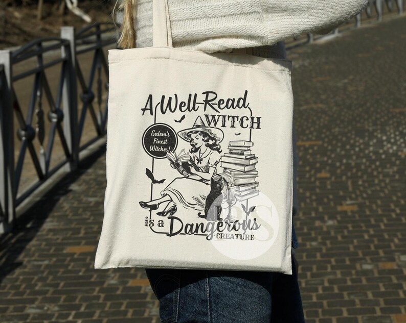 K&ouml;nnte beinhalten: Eine wei&szlig;e Einkaufstasche mit einer Schwarzwei&szlig;illustration einer Hexe, die ein Buch liest. Der Text auf der Tasche lautet "A Well-Read Witch" und "Salem's Finest Witches!" mit einem Stapel B&uuml;cher und dem Text "is a Dangerous Creature".