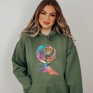 May include: Olive green hoodie with a colorful graphic of a disc golf basket and discs. The graphic has a watercolor effect with the text "Just Rip It". The hoodie has a drawstring hood and a front pocket.