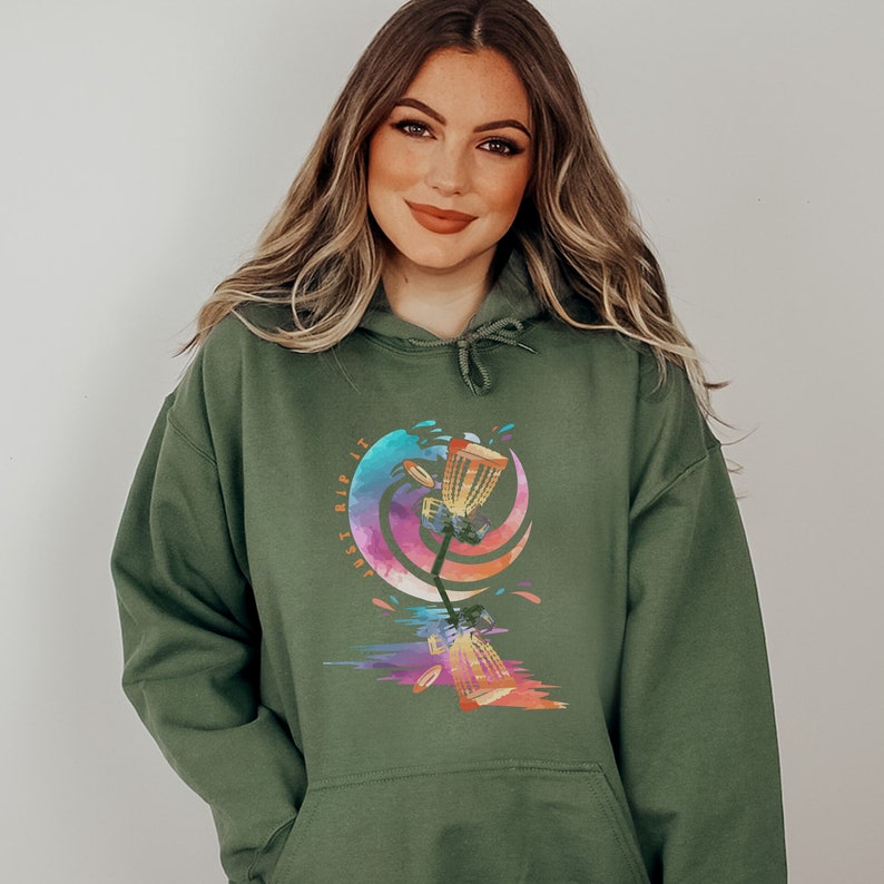 May include: Olive green hoodie with a colorful graphic of a disc golf basket and discs. The graphic has a watercolor effect with the text "Just Rip It". The hoodie has a drawstring hood and a front pocket.