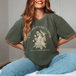 May include: A green t-shirt featuring a graphic print of a moon, flowers, and two figures. The text "A Midsummer Night's Dream" is printed in a circle around the graphic. The text "William Shakespeare" is printed below the graphic.
