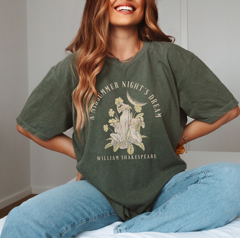 May include: A green t-shirt featuring a graphic print of a moon, flowers, and two figures. The text "A Midsummer Night's Dream" is printed in a circle around the graphic. The text "William Shakespeare" is printed below the graphic.
