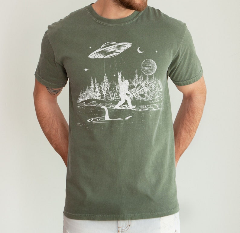 May include: A green t-shirt featuring a white graphic of a Bigfoot figure with a raised fist, standing in front of a lake with a Loch Ness Monster. A UFO hovers above Bigfoot, with stars and a crescent moon in the background.
