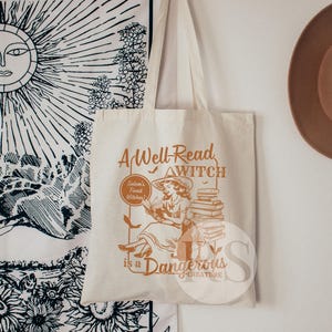 K&ouml;nnte beinhalten: Eine wei&szlig;e Canvas-Tragetasche mit einer braunen Grafik einer Hexe, die einen Stapel B&uuml;cher liest. Der Text auf der Tasche lautet "A Well-Read Witch is a Dangerous Creature".