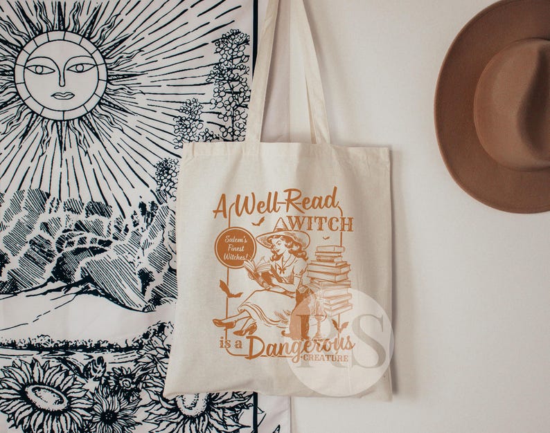 K&ouml;nnte beinhalten: Eine wei&szlig;e Canvas-Tragetasche mit einer braunen Grafik einer Hexe, die einen Stapel B&uuml;cher liest. Der Text auf der Tasche lautet "A Well-Read Witch is a Dangerous Creature".