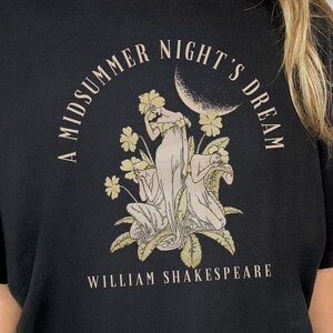May include: A black t-shirt featuring a white graphic print of three women in a garden setting. The text "A Midsummer Night's Dream" is printed in a circle around the women. The text "William Shakespeare" is printed below the graphic.