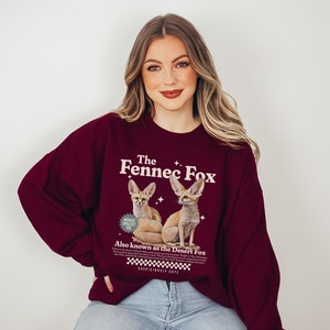 May include: A maroon sweatshirt with a graphic of two fennec foxes. The text on the sweatshirt reads "The Fennec Fox" and "Also known as the Desert Fox".