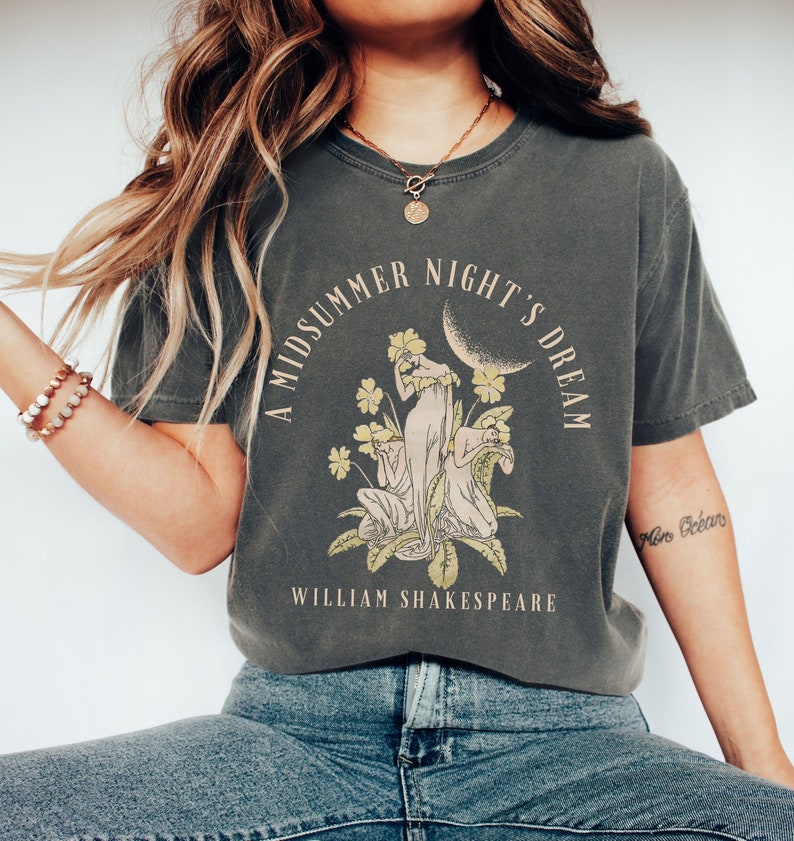 May include: A dark grey t-shirt with a graphic print of a woman with flowers and a crescent moon. The text "A Midsummer Night's Dream" is printed in a light brown font above the graphic. The text "William Shakespeare" is printed in a light brown font below the graphic.