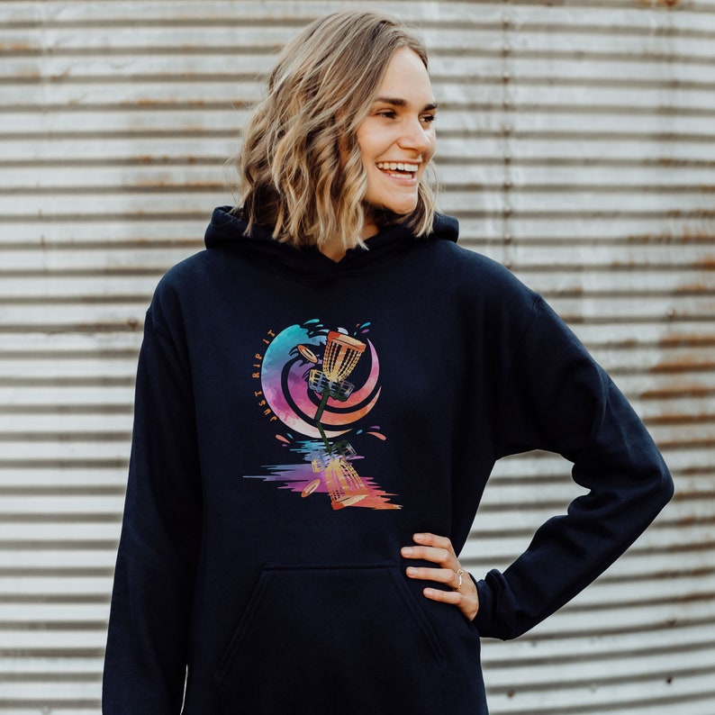 May include: Navy blue hoodie with a colorful graphic of a disc golf basket and the text "Just Rip It". The design features a vibrant watercolor style with shades of blue, pink, and orange. The hoodie has a front pocket and a hood.