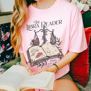 Pu&ograve; includere: Una maglietta rosa con un design grafico di un libro con una bilancia e il testo "The Libra Reader: Finding Beauty & Balance on Earth".