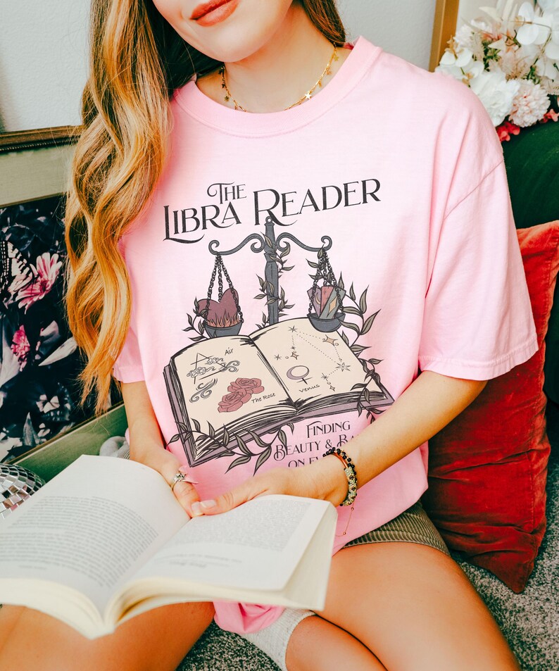 Pu&ograve; includere: Una maglietta rosa con un design grafico di un libro con una bilancia e il testo "The Libra Reader: Finding Beauty & Balance on Earth".