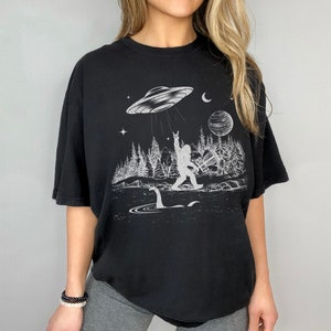 May include: Black t-shirt featuring a white graphic print of a Bigfoot character holding up a rock and roll hand gesture. The graphic also includes a UFO, a moon, a planet, and a Loch Ness Monster.