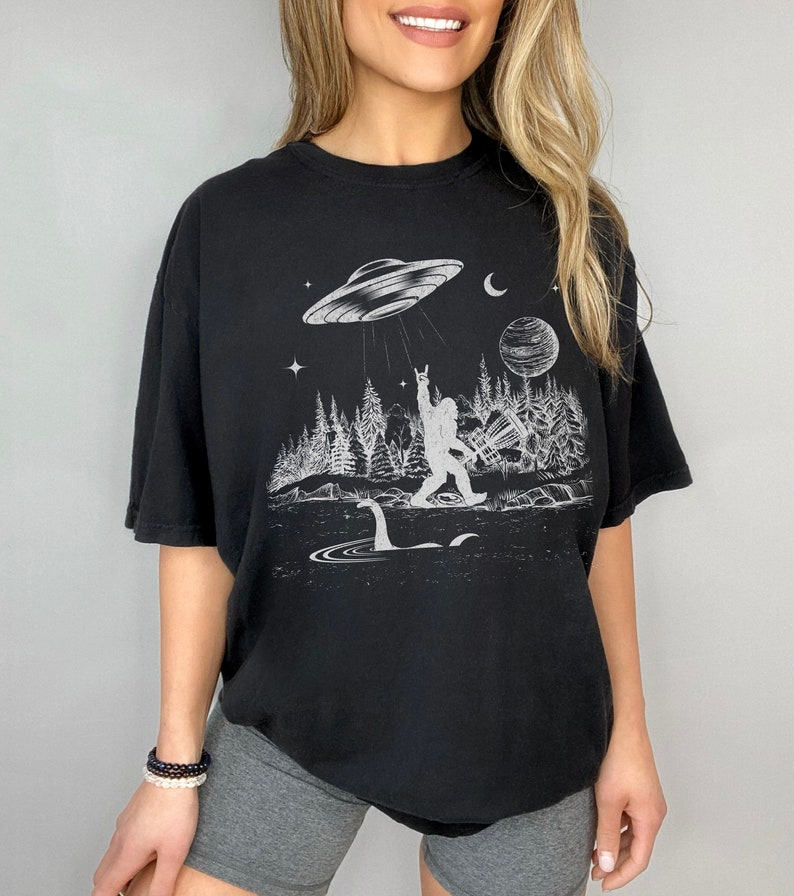 May include: Black t-shirt featuring a white graphic print of a Bigfoot character holding up a rock and roll hand gesture. The graphic also includes a UFO, a moon, a planet, and a Loch Ness Monster.