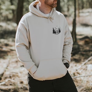 May include: A beige hoodie with a black graphic of a disc golf basket in a forest setting. The graphic is on the left chest of the hoodie.