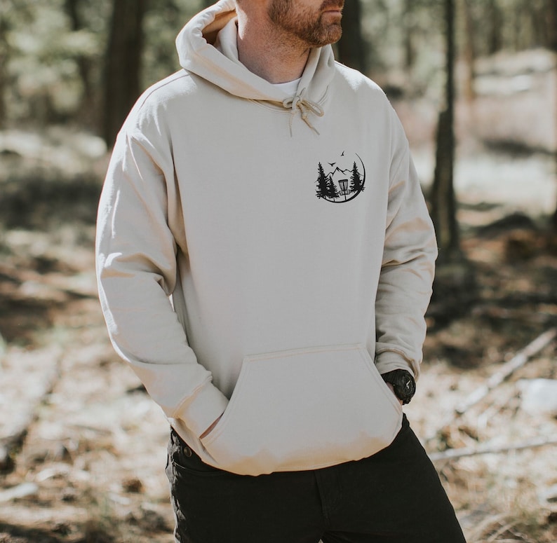 May include: A beige hoodie with a black graphic of a disc golf basket in a forest setting. The graphic is on the left chest of the hoodie.