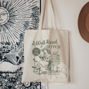K&ouml;nnte beinhalten: Eine wei&szlig;e Canvas-Tragetasche mit einer gr&uuml;nen Grafik einer Frau, die ein Buch liest. Der Text auf der Tasche lautet "A Well-Read Witch is a Dangerous Creature".