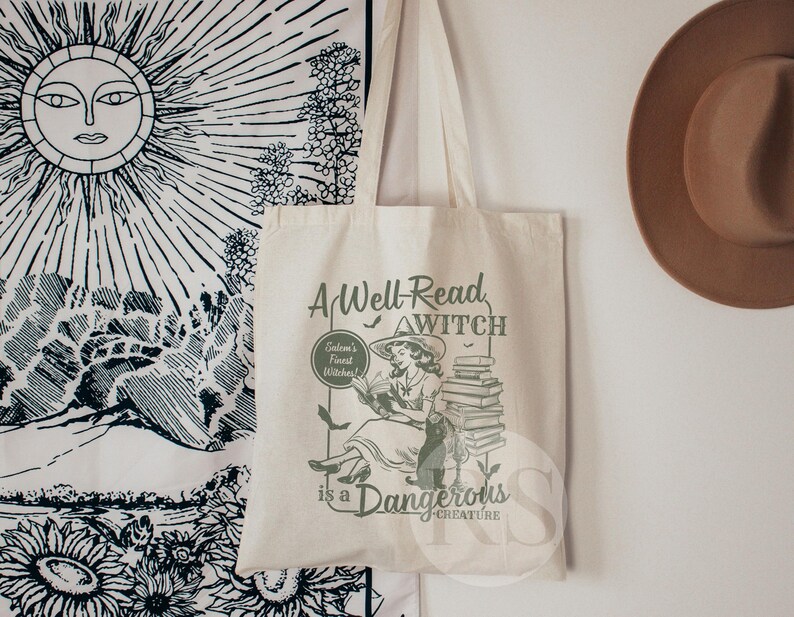 K&ouml;nnte beinhalten: Eine wei&szlig;e Canvas-Tragetasche mit einer gr&uuml;nen Grafik einer Frau, die ein Buch liest. Der Text auf der Tasche lautet "A Well-Read Witch is a Dangerous Creature".