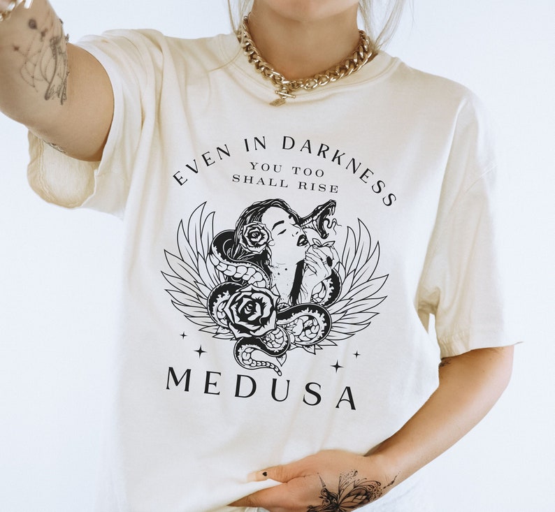 May include: Cream-colored t-shirt featuring a black and white graphic of Medusa with snakes, roses, and wings. The text "EVEN IN DARKNESS YOU TOO SHALL RISE" and "MEDUSA" are printed above and below the graphic. The shirt has short sleeves.