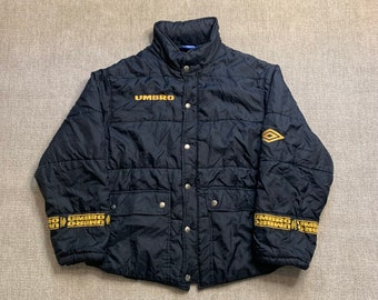 May include: A black quilted jacket with a gold "UMBRO" logo on the front and a diamond logo on the right sleeve. The jacket has a snap closure and two pockets on the front. The cuffs and waistband have a gold "UMBRO" logo.