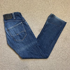 May include: A pair of blue denim jeans with a dark blue patch on the right leg and a small leather patch with the letters "GS" on the left side of the waistband.