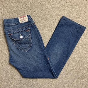 Könnte beinhalten: Eine blaue Jeans aus Denim mit roten Nähten an den Gesäßtaschen. Die Jeans hat einen kleinen, weißen Knopf an der Gesäßtasche und ein True Religion-Etikett am Bund.