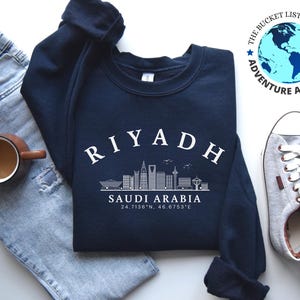Op de afbeelding: Een marineblauw sweatshirt met een witte afbeelding van de skyline van Riyad en de tekst "RIYADH" boven de skyline. De tekst "SAUDI-ARABIË" staat onder de skyline, samen met de coördinaten "24.7136°N, 46.6753°E".