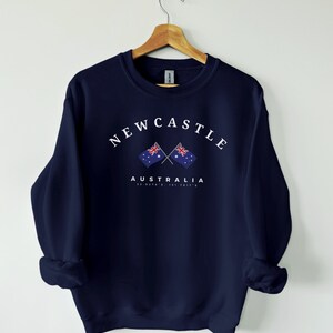 May include: Navy blue sweatshirt with a white graphic of two crossed Australian flags and the text "NEWCASTLE AUSTRALIA 32.0278&deg; S, 151.7811&deg; E".