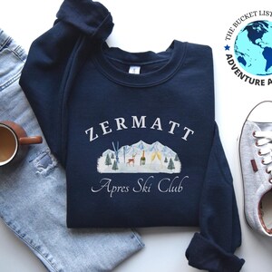 May include: Navy blue sweatshirt with the text "Zermatt Apres Ski Club" in white. The design includes a mountain scene with trees, a deer, and a tent. The sweatshirt is paired with light blue jeans, a coffee mug, and gray sneakers.