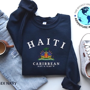 May include: A navy blue sweatshirt with the word "HAITI" in white arched lettering. Below is a colorful emblem and the word "CARIBBEAN" with coordinates. The sweatshirt is next to a pair of light blue jeans, a coffee mug, and gray sneakers.