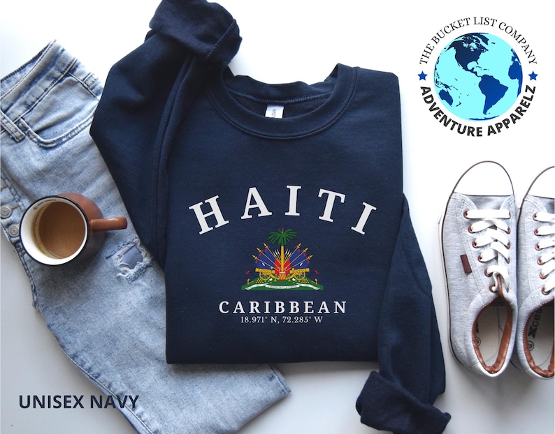 May include: A navy blue sweatshirt with the word "HAITI" in white arched lettering. Below is a colorful emblem and the word "CARIBBEAN" with coordinates. The sweatshirt is next to a pair of light blue jeans, a coffee mug, and gray sneakers.