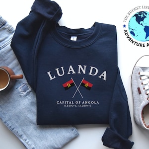 May include: A navy blue sweatshirt with the text "LUANDA" in white, "CAPITAL OF ANGOLA" in white, and the coordinates "8.8390°S, 13.2894°E" in white. Two crossed flags of Angola are shown below the text. The sweatshirt is laid out on a white surface with a pair of gray canvas shoes and a pair of blue jeans.