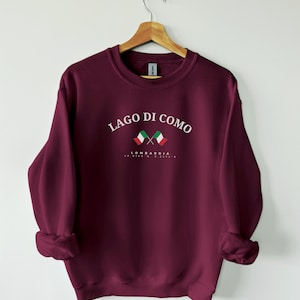 Puede incluir: Una sudadera burdeos con la inscripción blanca "LAGO DI COMO", acompañada de dos banderas italianas cruzadas. Debajo, "LOMBARDIA" y coordenadas. La sudadera está colgada de una percha de madera.