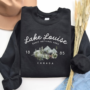 May include: Black sweatshirt with a white graphic design that says "Lake Louise Banff National Park 18 85 Canada". The graphic also features a watercolor illustration of mountains, trees, and a deer.