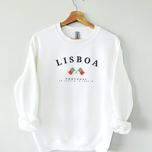 May include: White sweatshirt with black text that reads "LISBOA" and "PORTUGAL" with two crossed Portuguese flags and coordinates "38.7223&deg; N, 9.1393&deg; W".