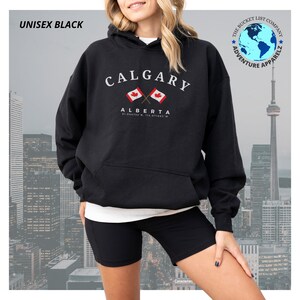 Pode incluir: Camisola de capuz unissexo preta com uma estampa gráfica branca do texto "CALGARY" com duas bandeiras canadenses cruzadas e o texto "ALBERTA" abaixo. O texto "51.044733', 114.071916'" está impresso abaixo do texto "ALBERTA".