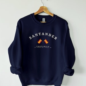 May include: Navy blue sweatshirt with the word "SANTANDER" in white arched text. Below the text are two crossed Spanish flags and the text "CANTABRIA" and coordinates. The sweatshirt is hanging on a wooden hanger.