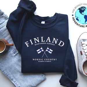 May include: A navy blue sweatshirt with the text "FINLAND" in white, "NORTHERN EUROPE" in white, and "NORDIC COUNTRY" in white. Two crossed Finnish flags are below the text. The coordinates "60.1695"N, 24.9354°E" are printed below the flags.