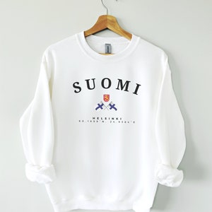 以下が含まれることがあります： 白いスウェットシャツに、黒い文字で「SUOMI」と書いてあり、2つの交差したフィンランドの国旗とフィンランドの国章のグラフィック、そして黒い文字で「HELSINKI 60.1699&deg; N, 24.9384&deg; E」と書いてあります。