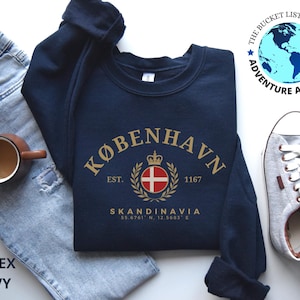 May include: Navy blue sweatshirt with gold lettering that reads "KØBENHAVN" and "SKANDINAVIA". The design includes a crest with a cross and crown. The text "UNISEX NAVY" is at the bottom left. A coffee mug, jeans, and sneakers are also in the image.