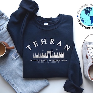 Pode incluir: Uma camisola azul marinho com a palavra "TEHRAN" impressa em branco na frente. A camisola também apresenta um horizonte da cidade com o texto "MÉDIO ORIENTE - ÁSIA OCIDENTAL" e as coordenadas "35.6892° N, 51.3890° E" impressas abaixo do horizonte.